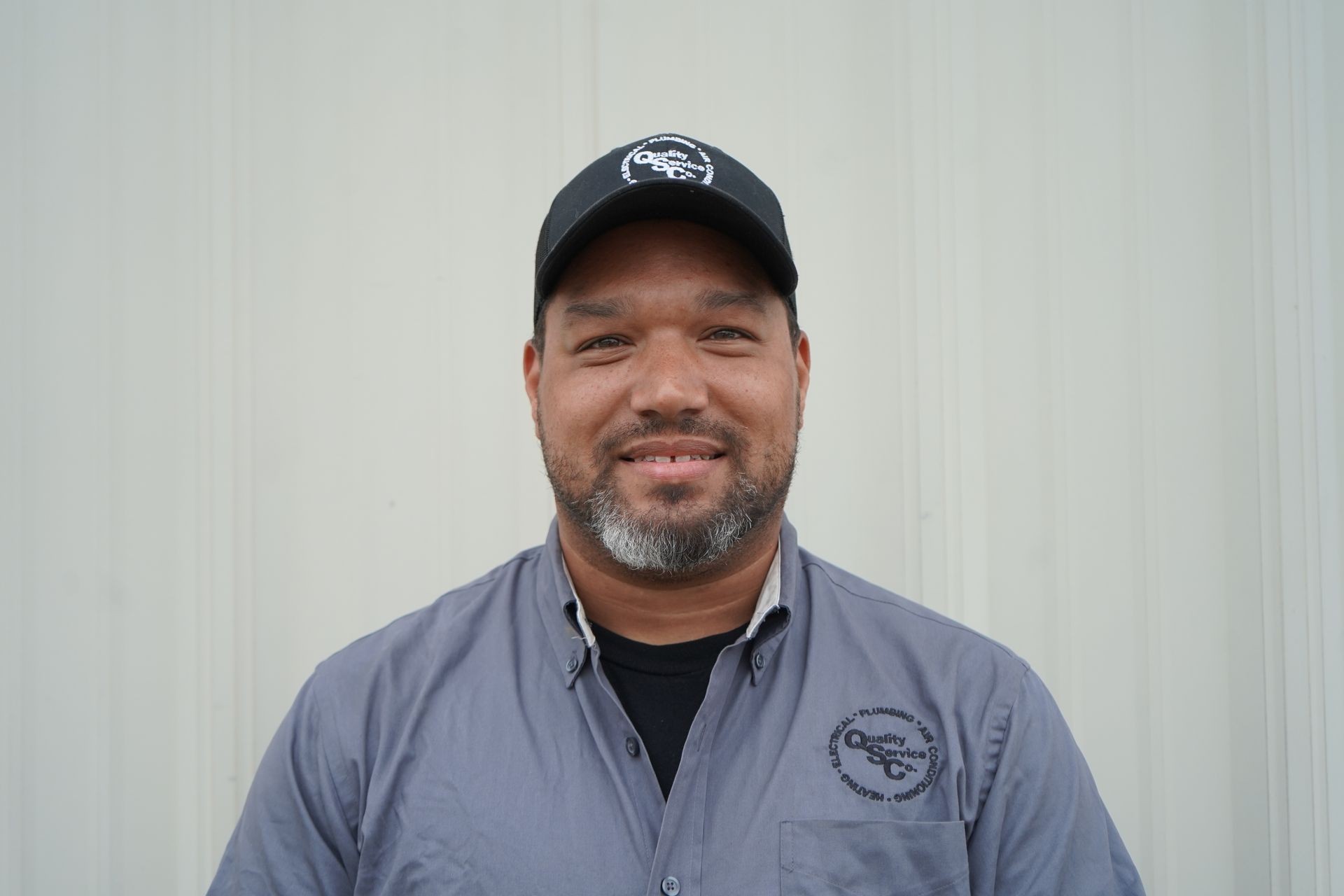 Smiling man wearing a black cap and gray shirt standing in front of a light background.
