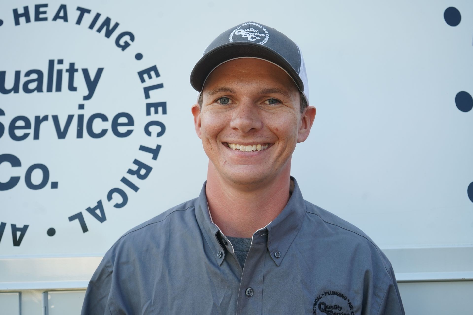Smiling person wearing a gray shirt and cap standing in front of a company logo background.