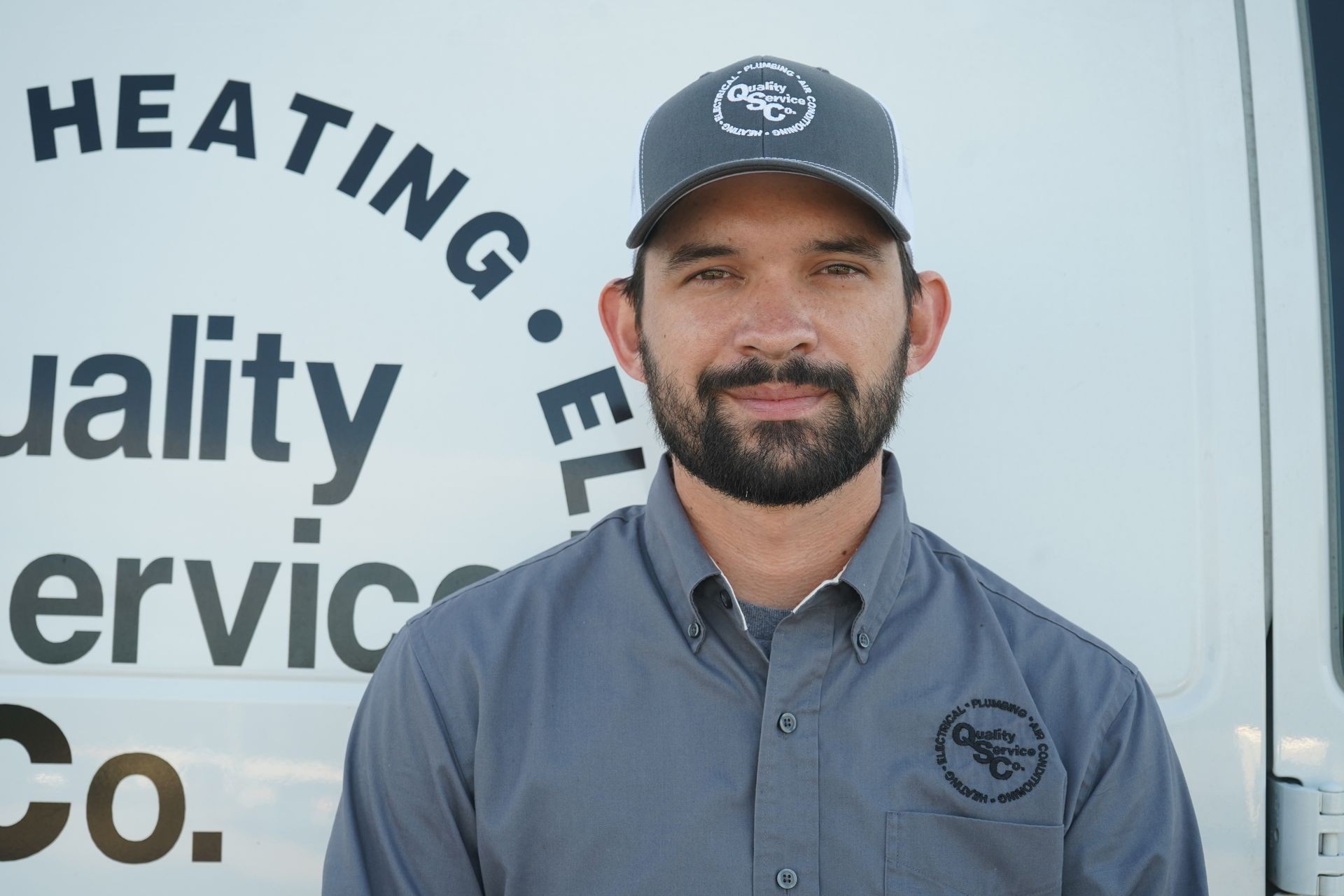 Man in a gray uniform and cap standing in front of a branded service vehicle with company logos.