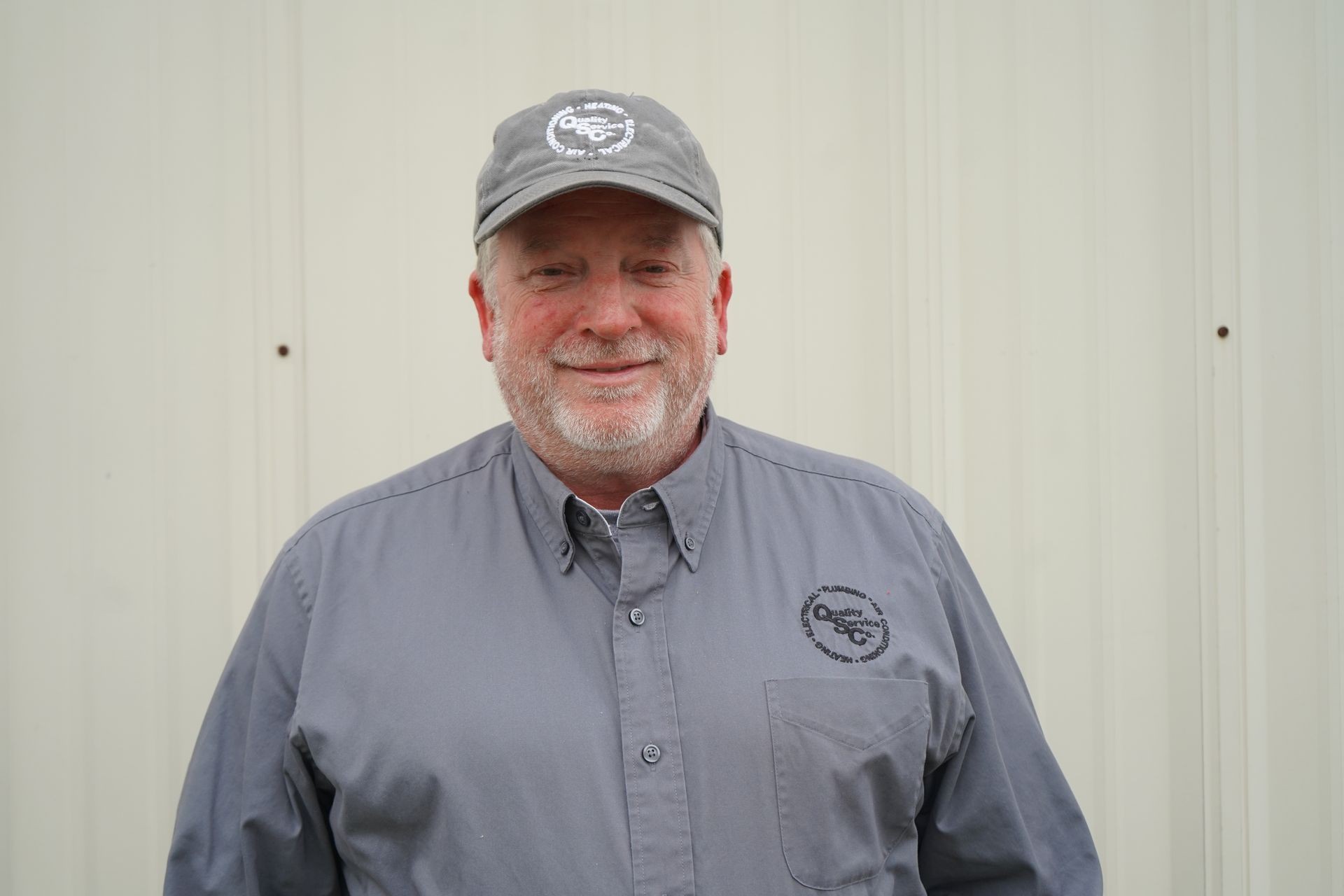 Person in a gray uniform and cap with company logo, standing against a light corrugated wall.