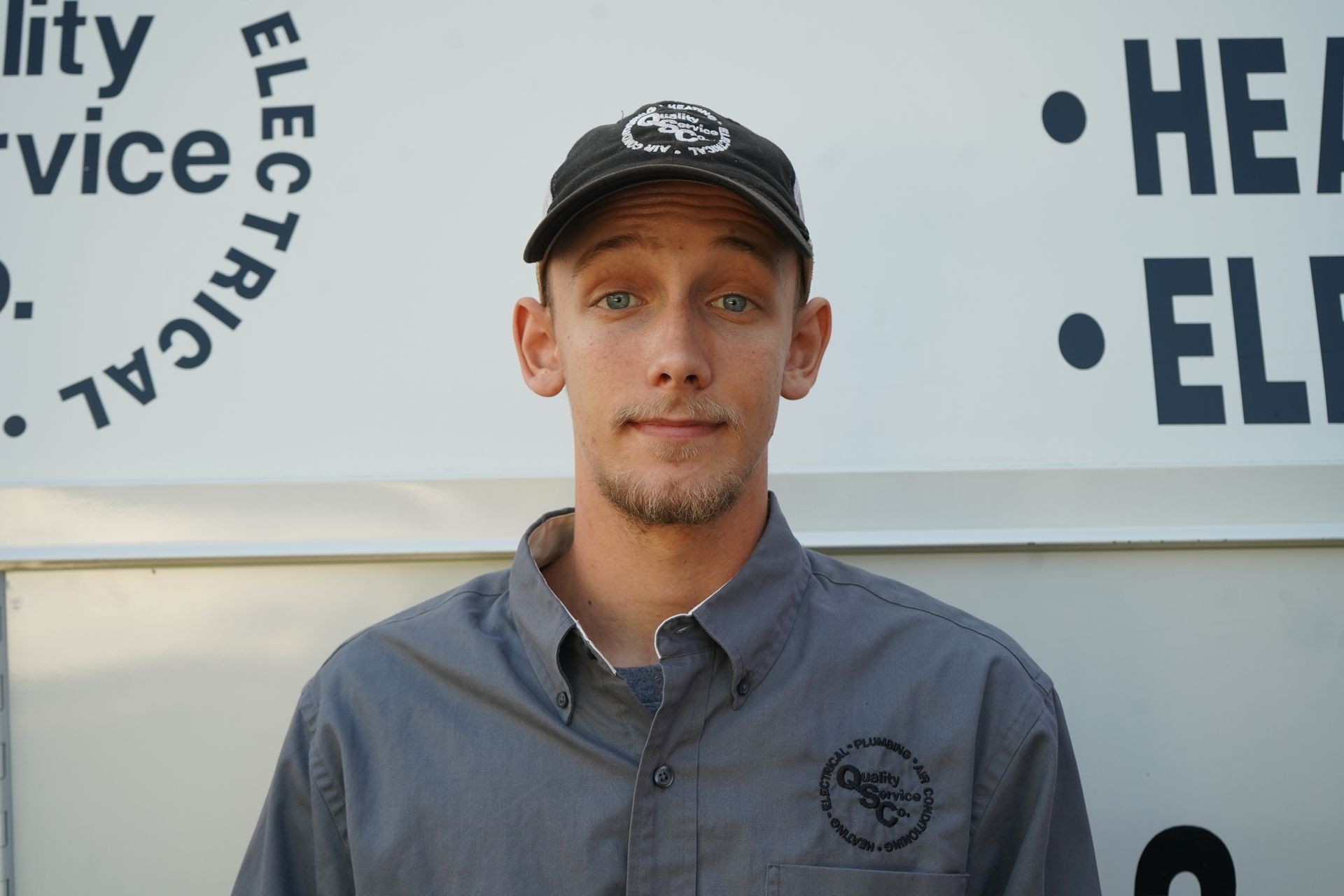 Person wearing a gray uniform and black cap standing in front of a company service vehicle.