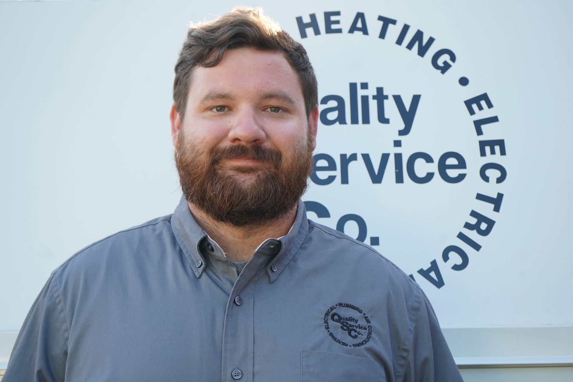 Man with a beard in a gray shirt, standing in front of a company logo wall.