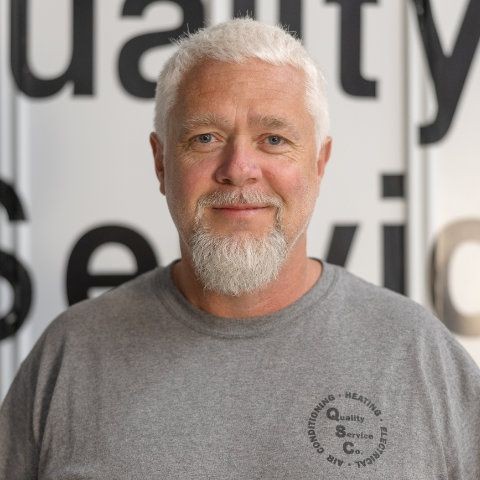 Man with a gray beard wearing a gray shirt in front of a blurred background with large letters.