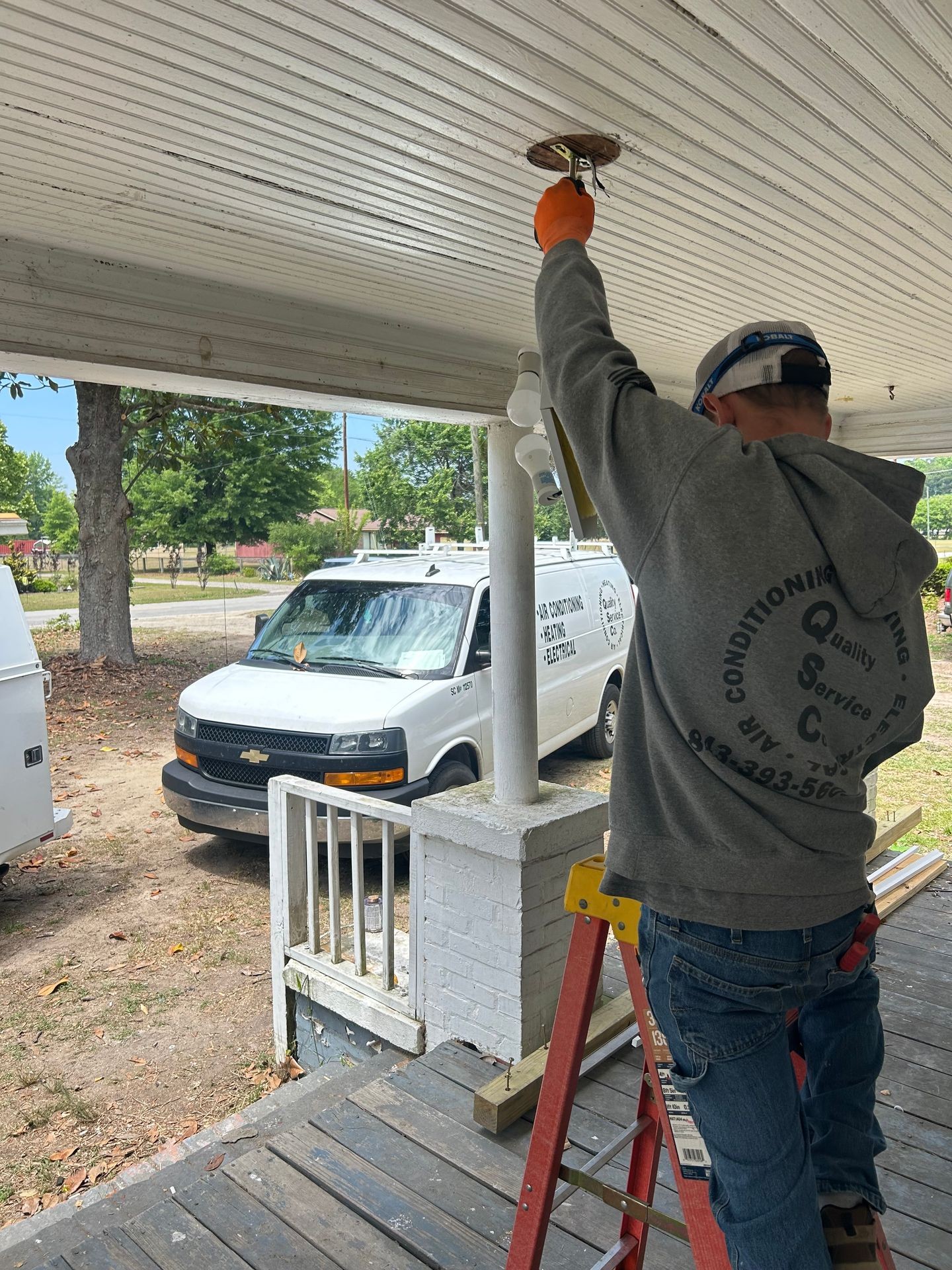 Person on a ladder fixing the ceiling on a porch, with a service van parked outside.