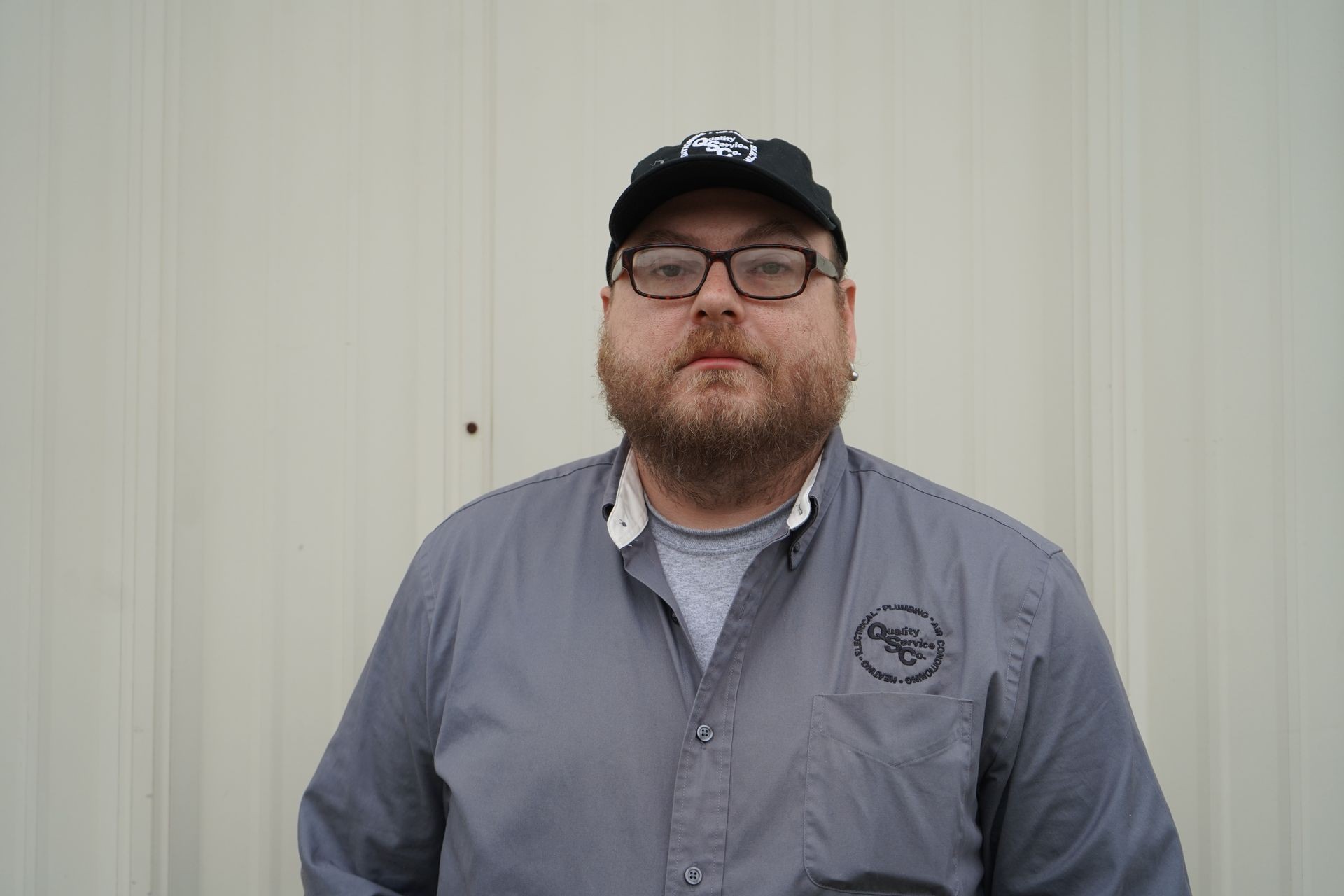 Man with glasses and a beard wearing a cap and gray shirt standing against a light-colored wall.