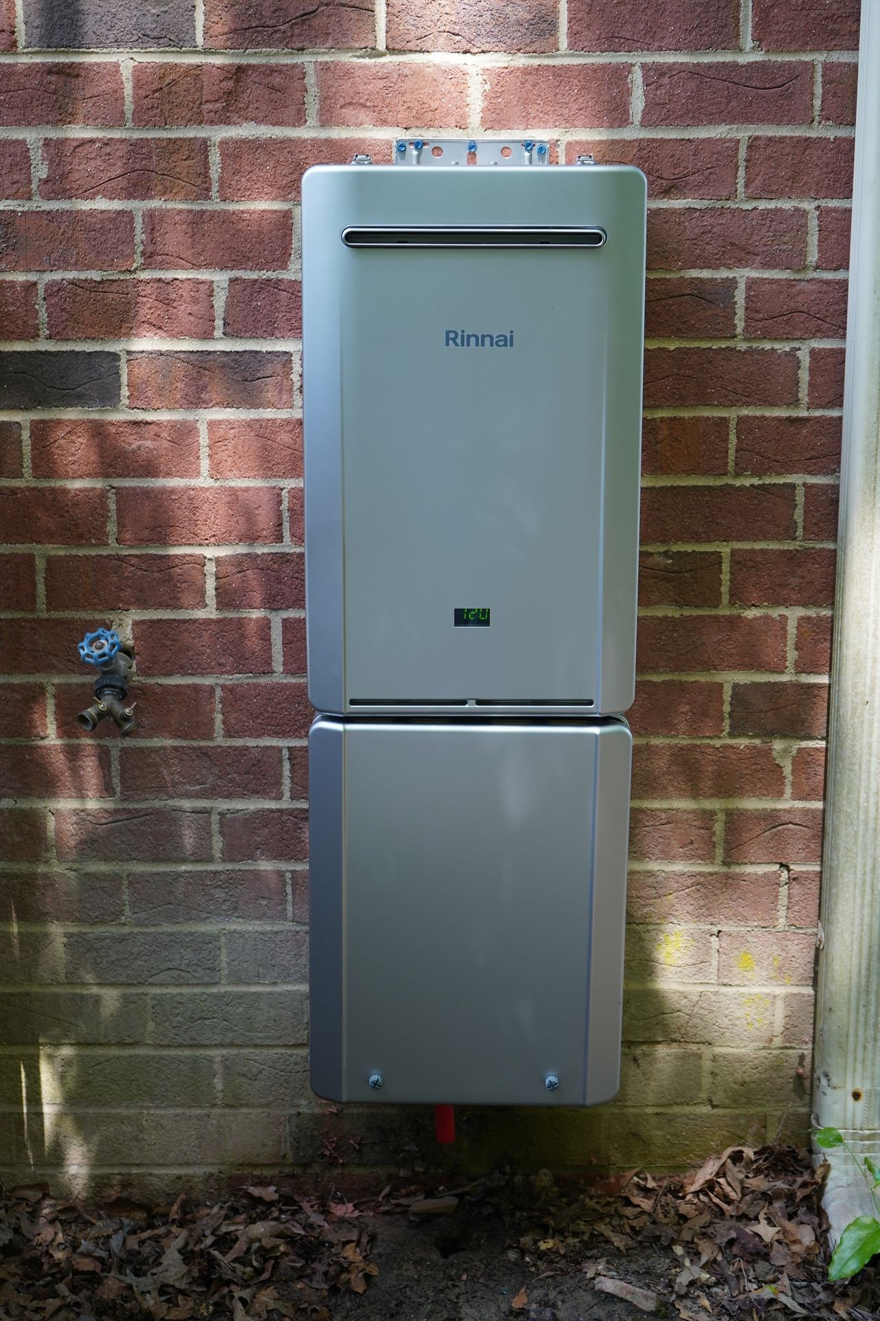 Outdoor gas water heater mounted on a brick wall with a garden faucet nearby.