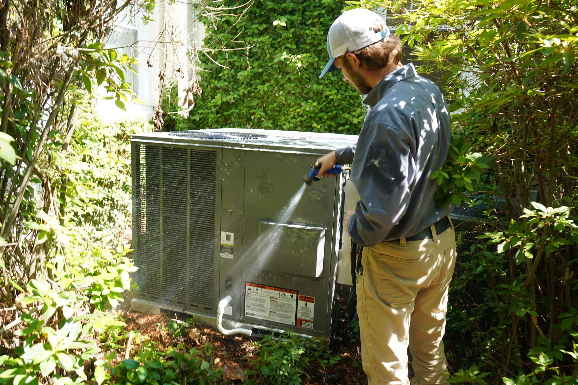 Person cleaning an outdoor air conditioning unit with a hose, surrounded by green foliage.